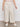 A person wears Sanctuary Clothing's soft marine wide leg crop pant macrame in cream, paired with a pink top and white sneakers, standing on a concrete floor against a plain white wall.