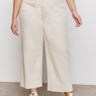A person wears Sanctuary Clothing's soft marine wide leg crop pant macrame in cream, paired with a pink top and white sneakers, standing on a concrete floor against a plain white wall.