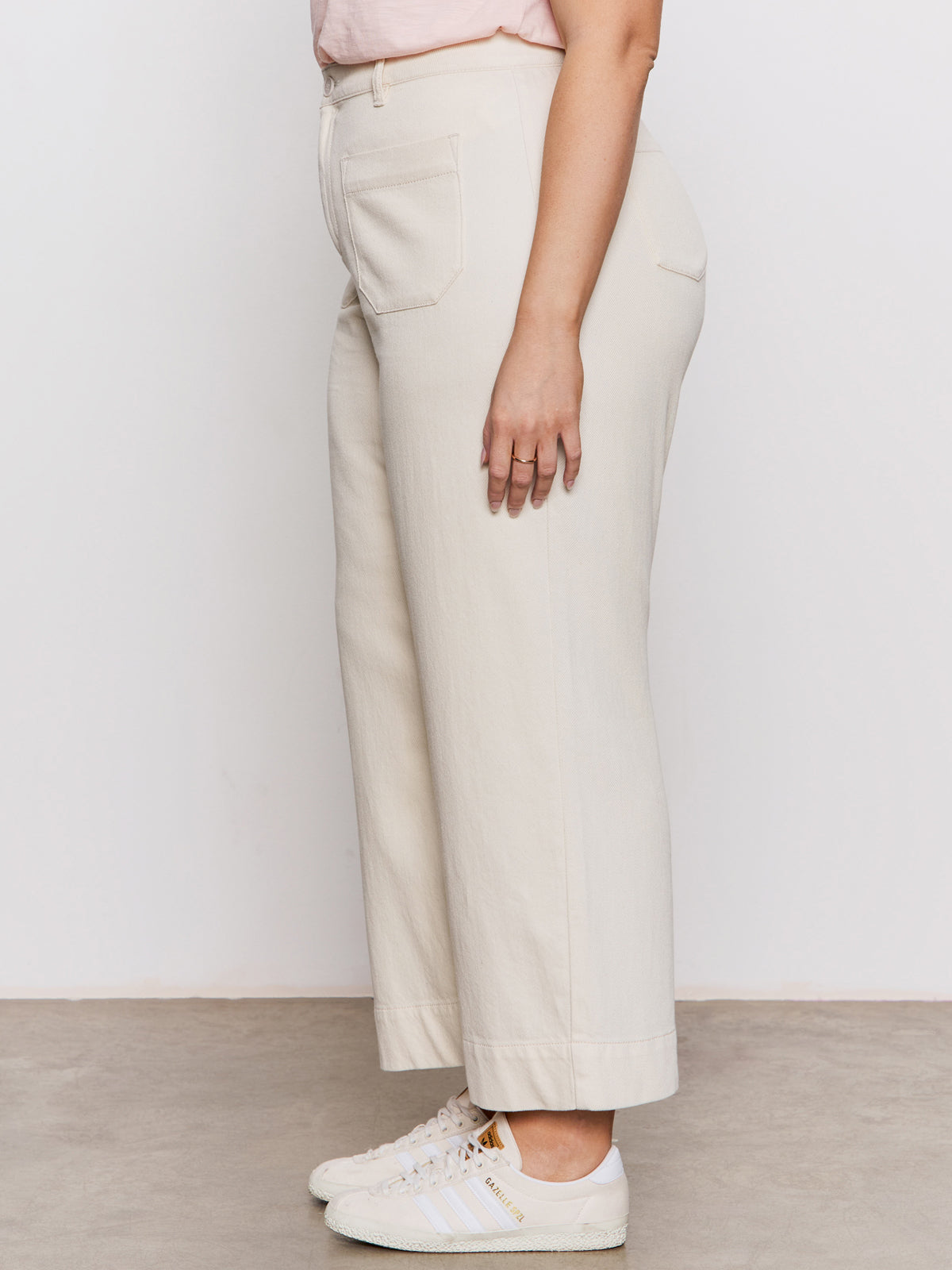 A person stands sideways in Sanctuary Clothing's soft marine wide leg crop pant macrame (extended sizing), off-white with front and back pockets, paired with white and beige sneakers against a neutral backdrop.