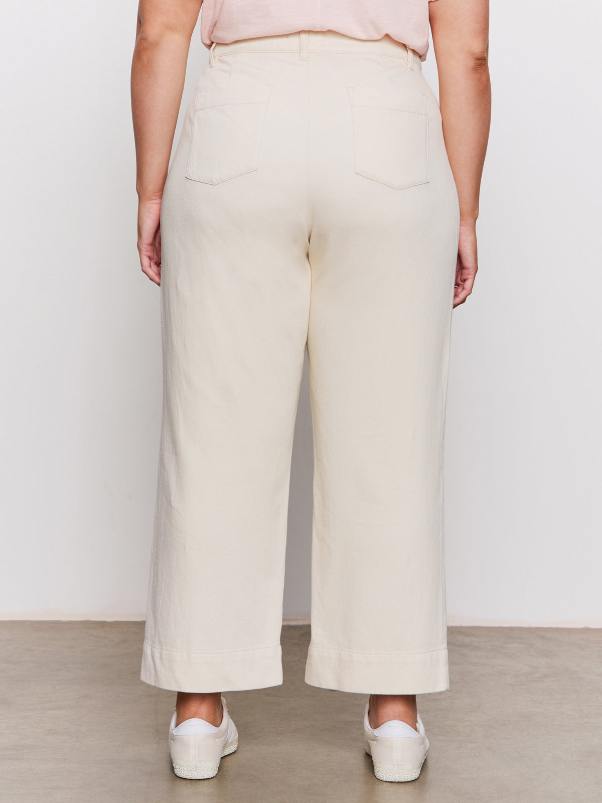 A person stands facing away, wearing high-waisted, wide-leg, off-white pants with two back pockets, a light pink top, and white shoes, against a plain background.