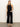 A woman with straight blond hair and a neutral expression stands against a plain wall, wearing a sleeveless black halter-neck top, Sanctuary Clothing soft marine wide leg crop pant in black, black heeled sandals, and earrings.