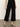 A person models the Sanctuary Clothing soft marine wide leg crop pant in black with black heeled sandals, standing on a concrete floor against a light background. The photo is cropped from the shoulders down.