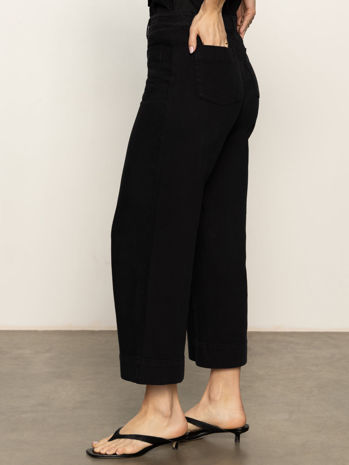 A person wearing the Sanctuary Clothing soft marine wide leg crop pant in black and black heeled sandals, with one hand on their hip, stands on a smooth indoor surface against a neutral background.