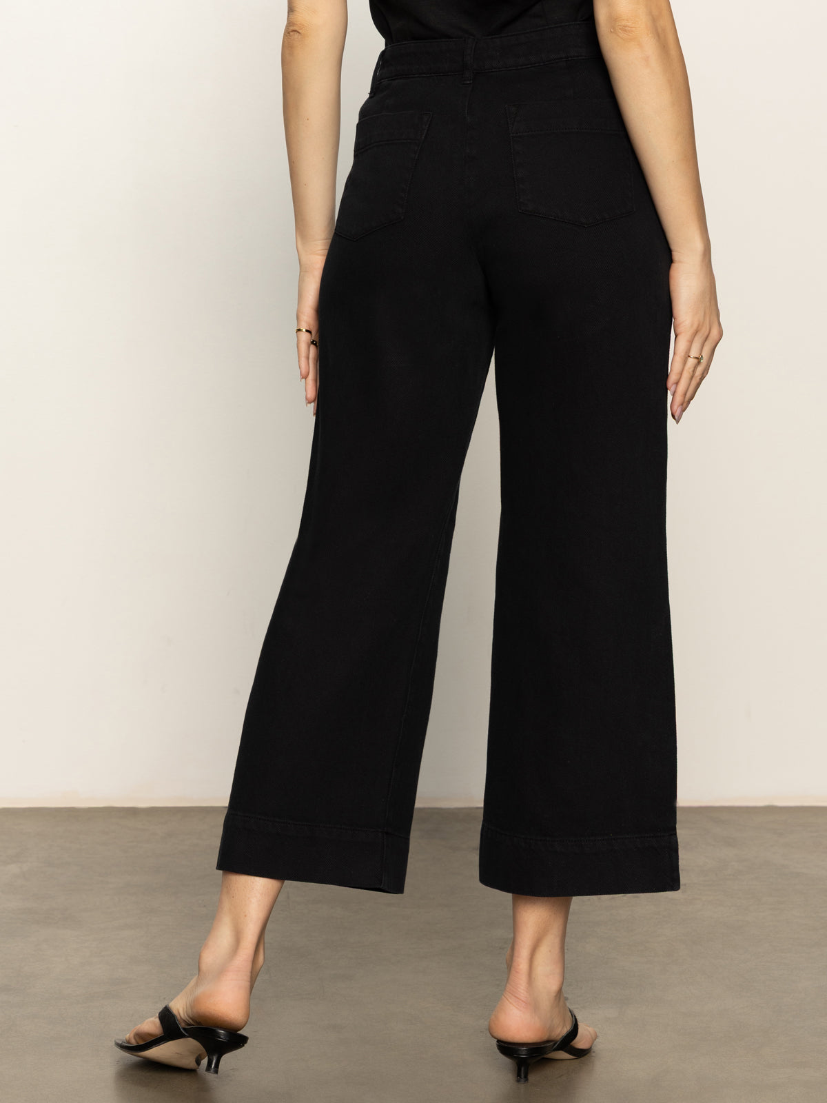 A person stands on a concrete floor, facing away in Sanctuary Clothing's soft marine wide leg crop pant in black with back pockets, paired with a black top and black heeled sandals; one hand is relaxed by their side.