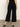 A person stands on a concrete floor, facing away in Sanctuary Clothing's soft marine wide leg crop pant in black with back pockets, paired with a black top and black heeled sandals; one hand is relaxed by their side.
