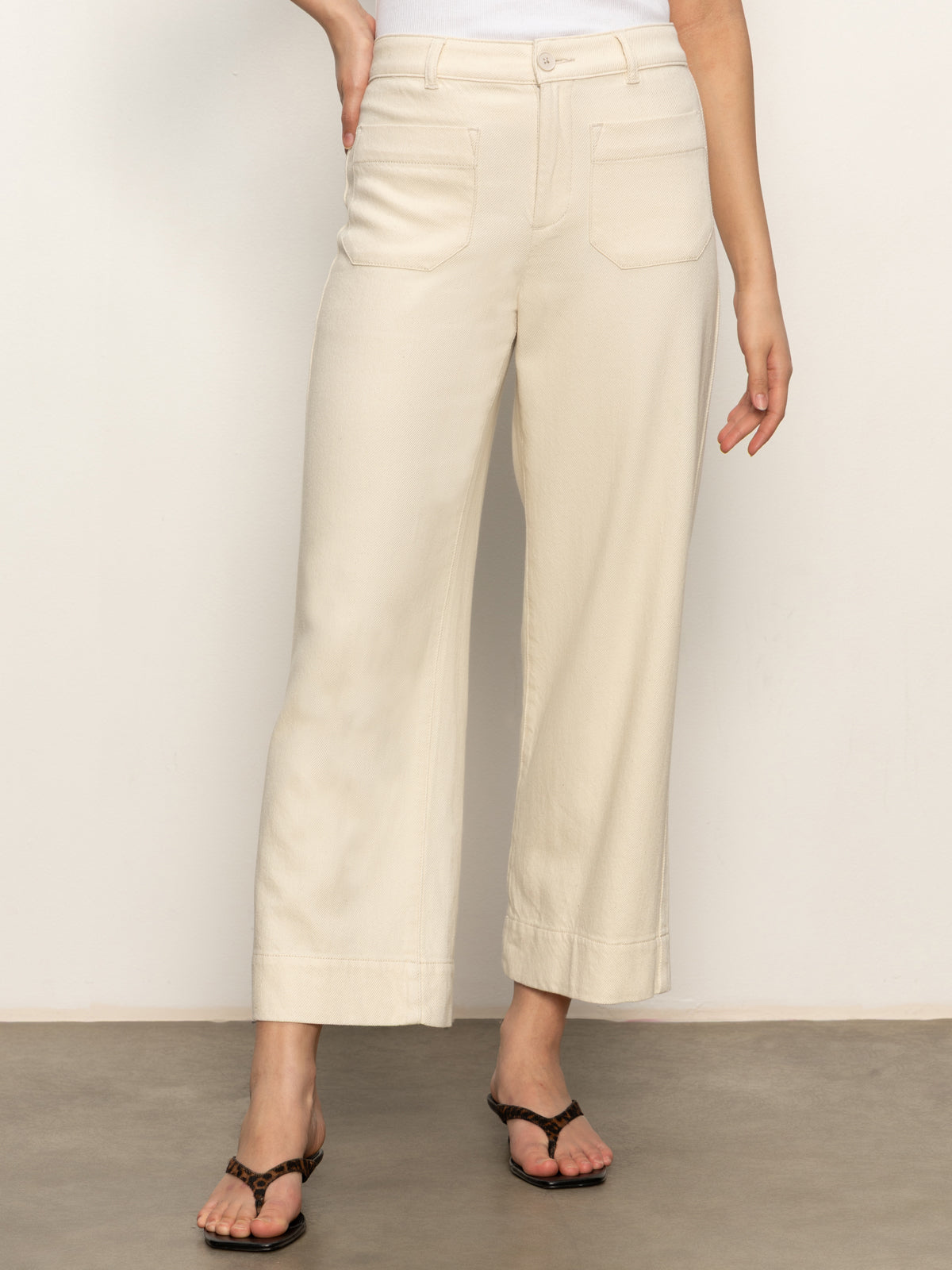 A person stands against a light background wearing the Sanctuary Clothing soft marine wide leg crop pant macrame in cream, featuring front patch pockets and black sandals. Their upper body is mostly out of frame.