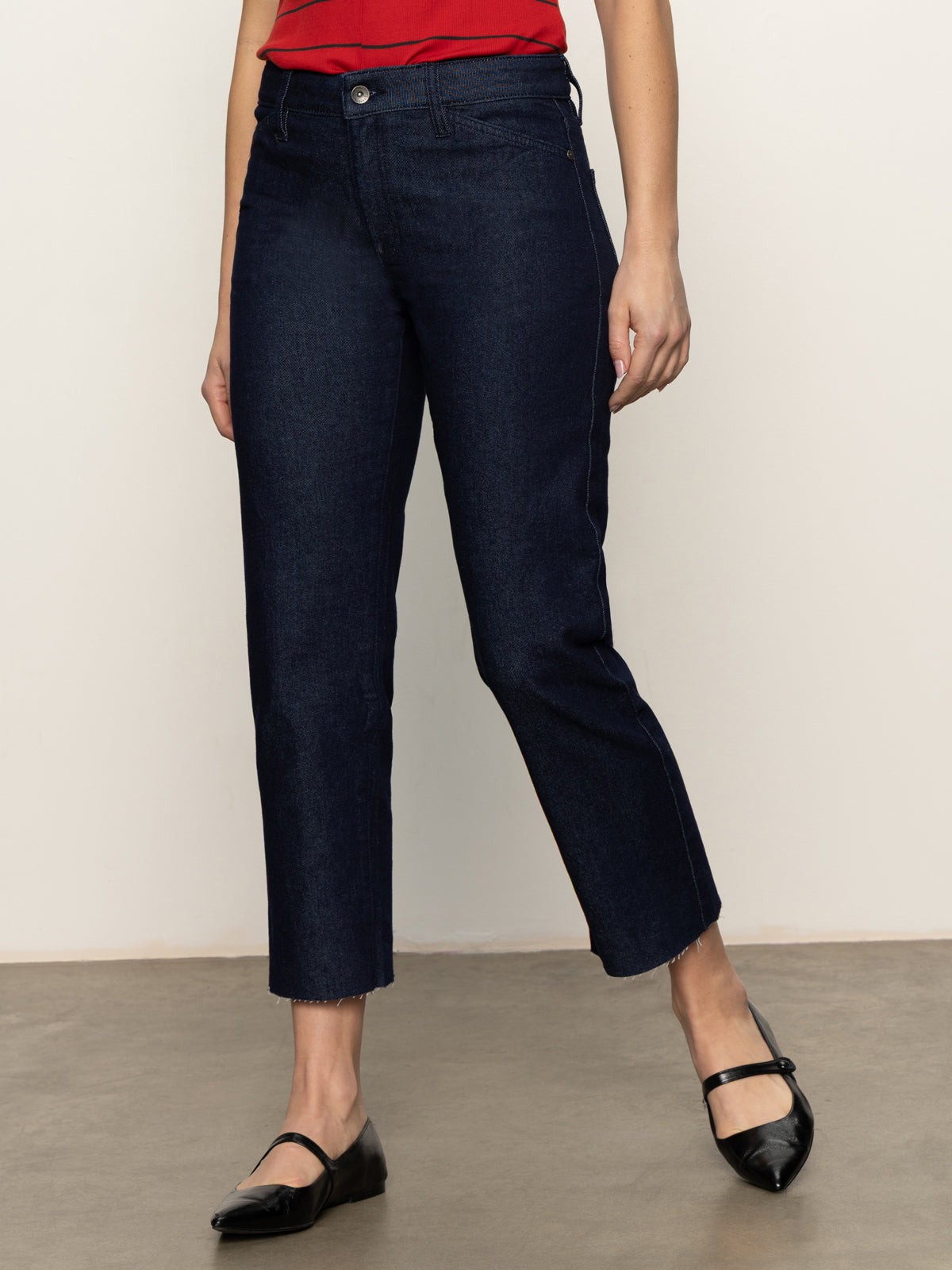A person wearing Sanctuary Clothing's "cadence 90s straight crop plot twist" jeans in dark blue, paired with a red top and black Mary Jane shoes, stands on a gray floor against a plain light background. Their face is not visible.
