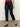 A person wearing Sanctuary Clothing's "cadence 90s straight crop plot twist" jeans in dark blue, paired with a red top and black Mary Jane shoes, stands on a gray floor against a plain light background. Their face is not visible.