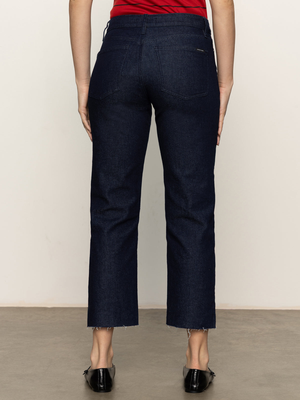 A person wearing Sanctuary Clothing's cadence 90s straight crop plot twist jeans, shown from the back with raw hems and black shoes against a plain light background.