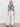 A woman with long blonde hair and a relaxed expression stands against a white wall wearing Sanctuary Clothing's cruiser full length powdered sugar pants, a green and white leaf tee, and black sandals.
