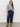 A woman poses against a white wall wearing Sanctuary Clothing's league crop plot twist extended sizing top, paired with dark blue high-waisted jeans and black sandals. One hand is in her pocket, the other by her side.