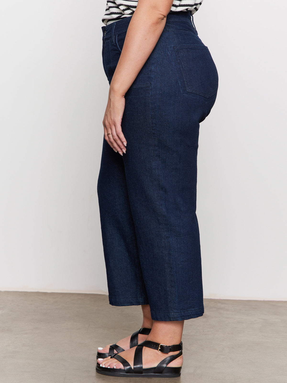 A woman stands sideways wearing Sanctuary Clothing’s league crop plot twist extended sizing top, high-waisted dark blue straight-leg jeans, and black platform sandals on a light floor against a plain wall.
