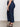 A woman stands sideways wearing Sanctuary Clothing’s league crop plot twist extended sizing top, high-waisted dark blue straight-leg jeans, and black platform sandals on a light floor against a plain wall.
