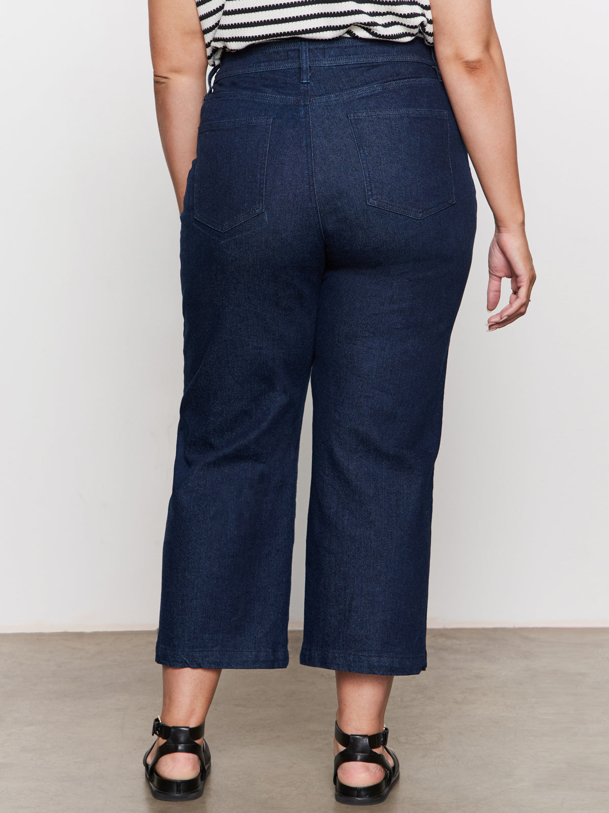 A person stands facing away, wearing Sanctuary Clothing’s league crop plot twist extended sizing: dark blue high-waisted, wide-leg jeans with back pockets, black sandals, and a black-and-white striped sleeveless top.