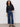 A woman with curly hair wears the "league crop plot twist" black blouse by Sanctuary Clothing, paired with blue wide-leg jeans and black sandals, standing against a plain light wall with one hand in her pocket.