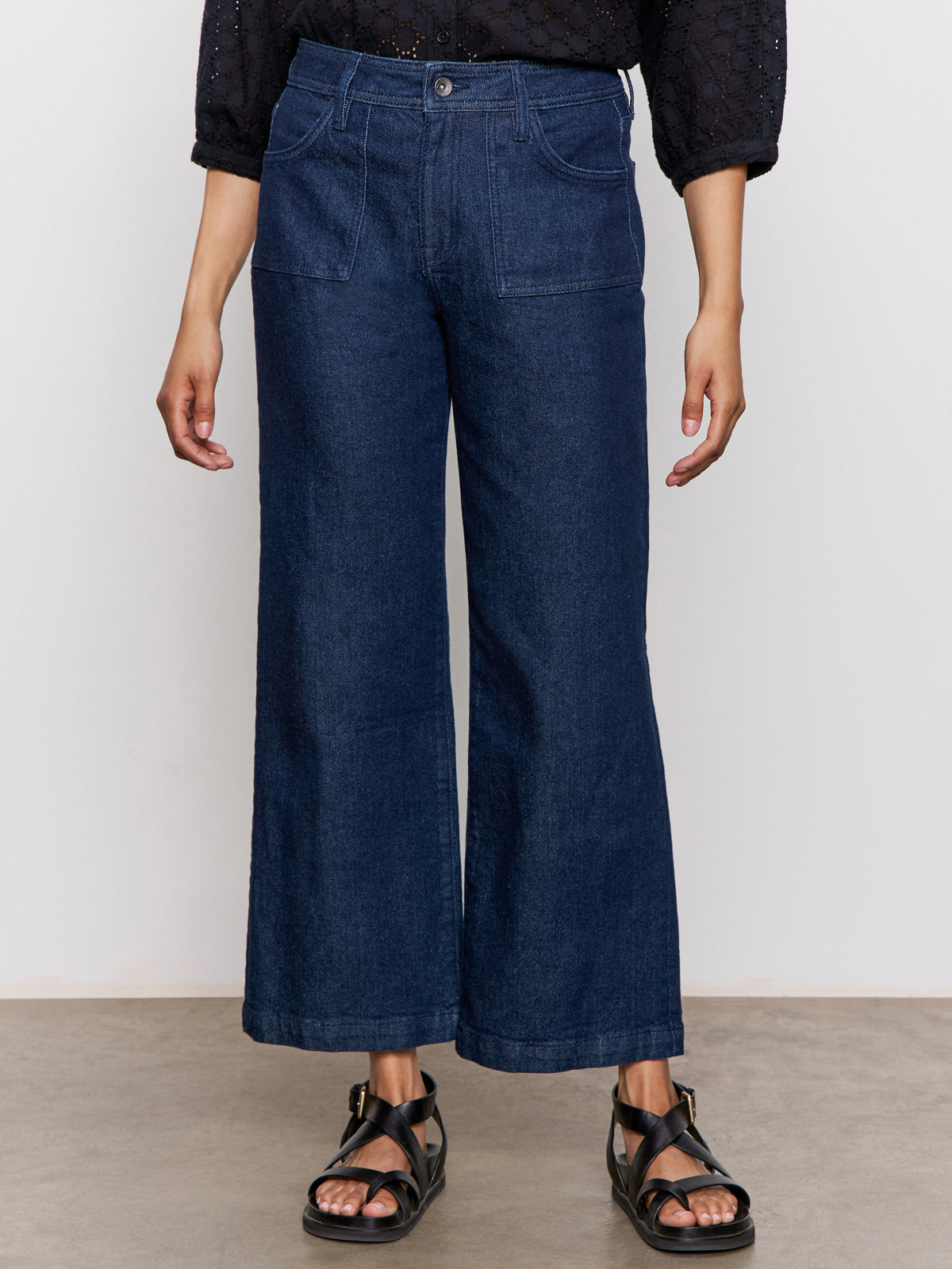 A person wears Sanctuary Clothing's league crop plot twist black textured long-sleeve shirt, paired with dark blue wide-leg jeans with front patch pockets and black strappy sandals, standing against a plain background.