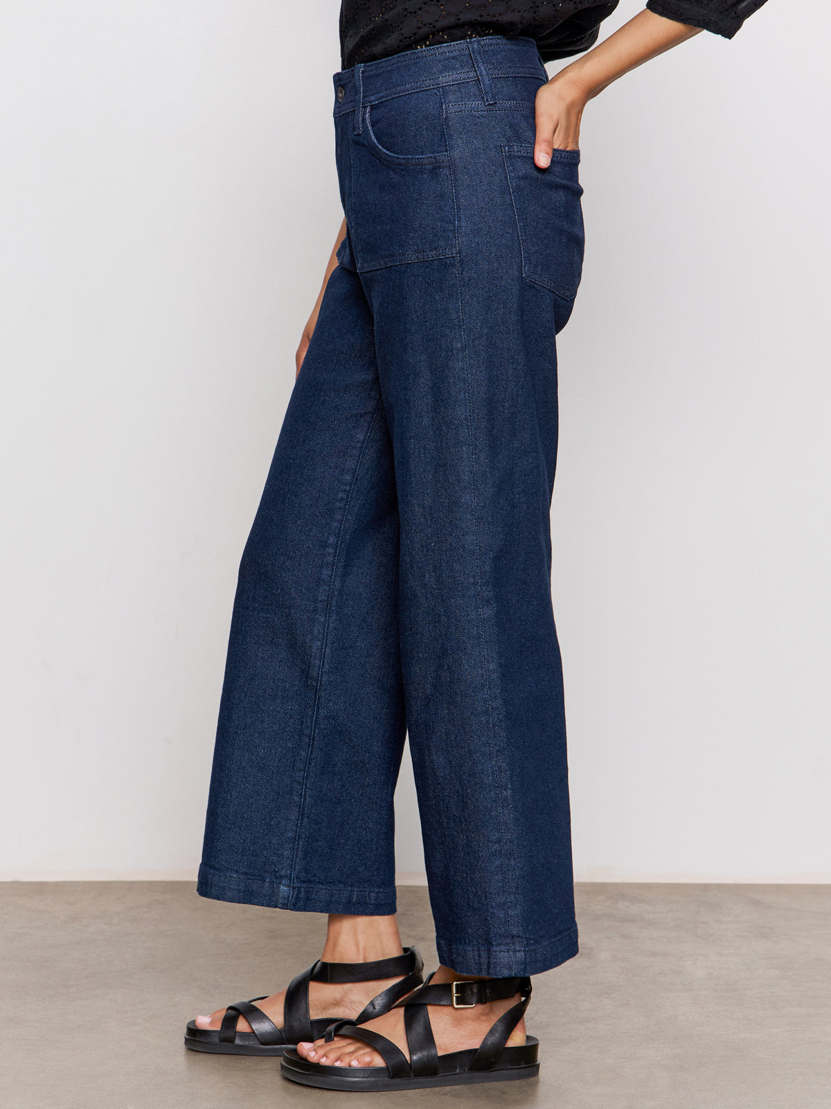 A person stands against a plain background wearing Sanctuary Clothing's league crop plot twist top, paired with wide-leg dark blue jeans and black sandals, with one hand in their back pocket.