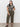 A woman with long dark hair and a neutral expression stands against a white wall wearing Sanctuary Clothing’s league crop fatigue pants in olive green, paired with a black and beige abstract print t-shirt and black sandals, one hand resting on her leg.