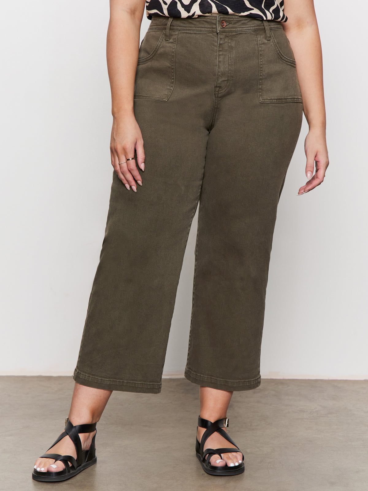 A person wearing Sanctuary Clothing's league crop fatigue extended sizing high-waisted olive green wide-leg pants with front pockets, paired with a black and beige patterned top and black strappy sandals, stands on a gray floor against a white wall.