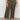 A person wearing Sanctuary Clothing's league crop fatigue extended sizing high-waisted olive green wide-leg pants with front pockets, paired with a black and beige patterned top and black strappy sandals, stands on a gray floor against a white wall.