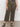 A person wearing Sanctuary Clothing's league crop fatigue extended sizing high-waisted olive green wide-leg pants with front pockets, paired with a black and beige patterned top and black strappy sandals, stands on a gray floor against a white wall.