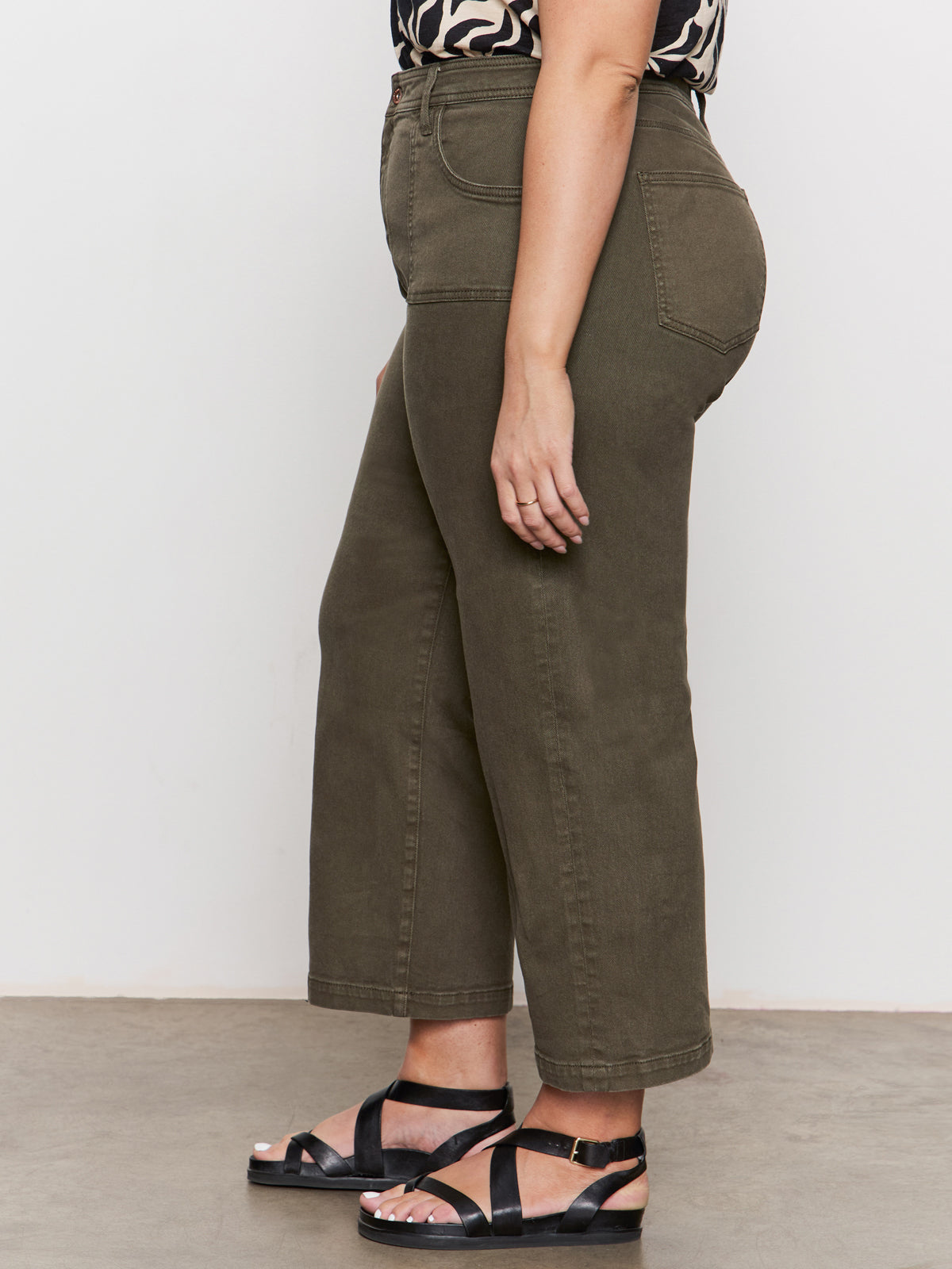 A person wears Sanctuary Clothing’s league crop fatigue extended sizing olive green high-waisted, straight-leg jeans, a patterned top, and black strappy sandals, standing sideways against a plain white background.