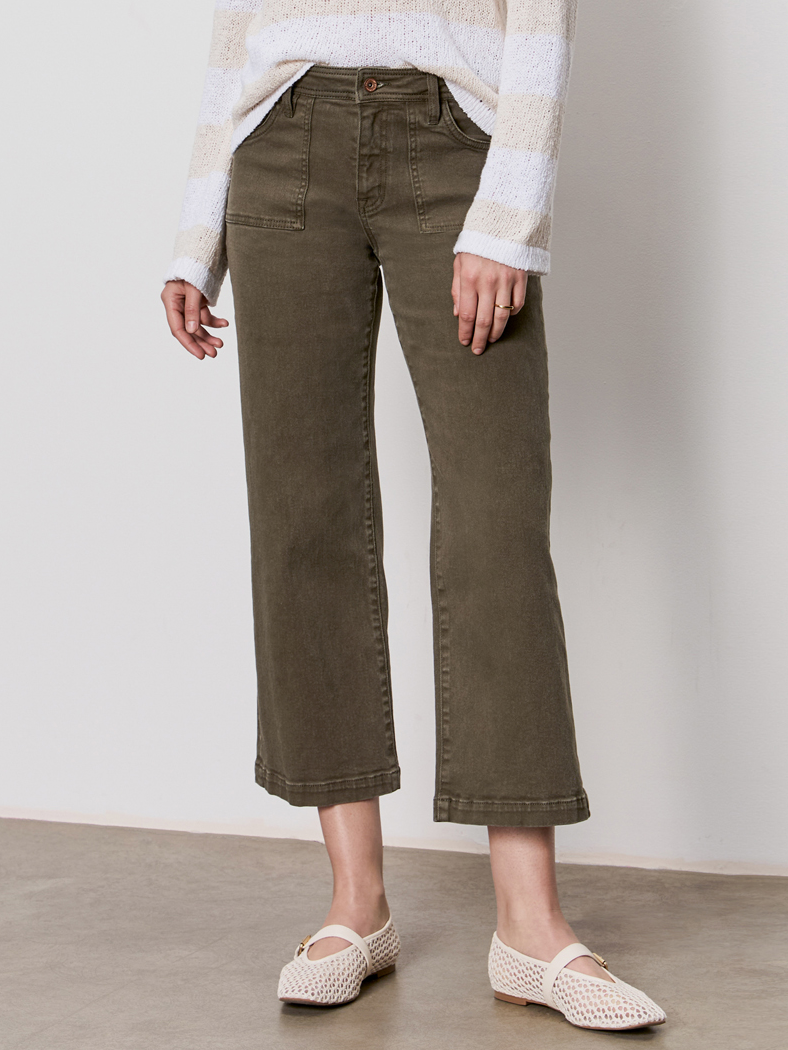 A person wearing Sanctuary Clothing's league crop fatigue pants, paired with a light long-sleeve sweater and white woven flats, stands indoors on a concrete floor against a plain wall.