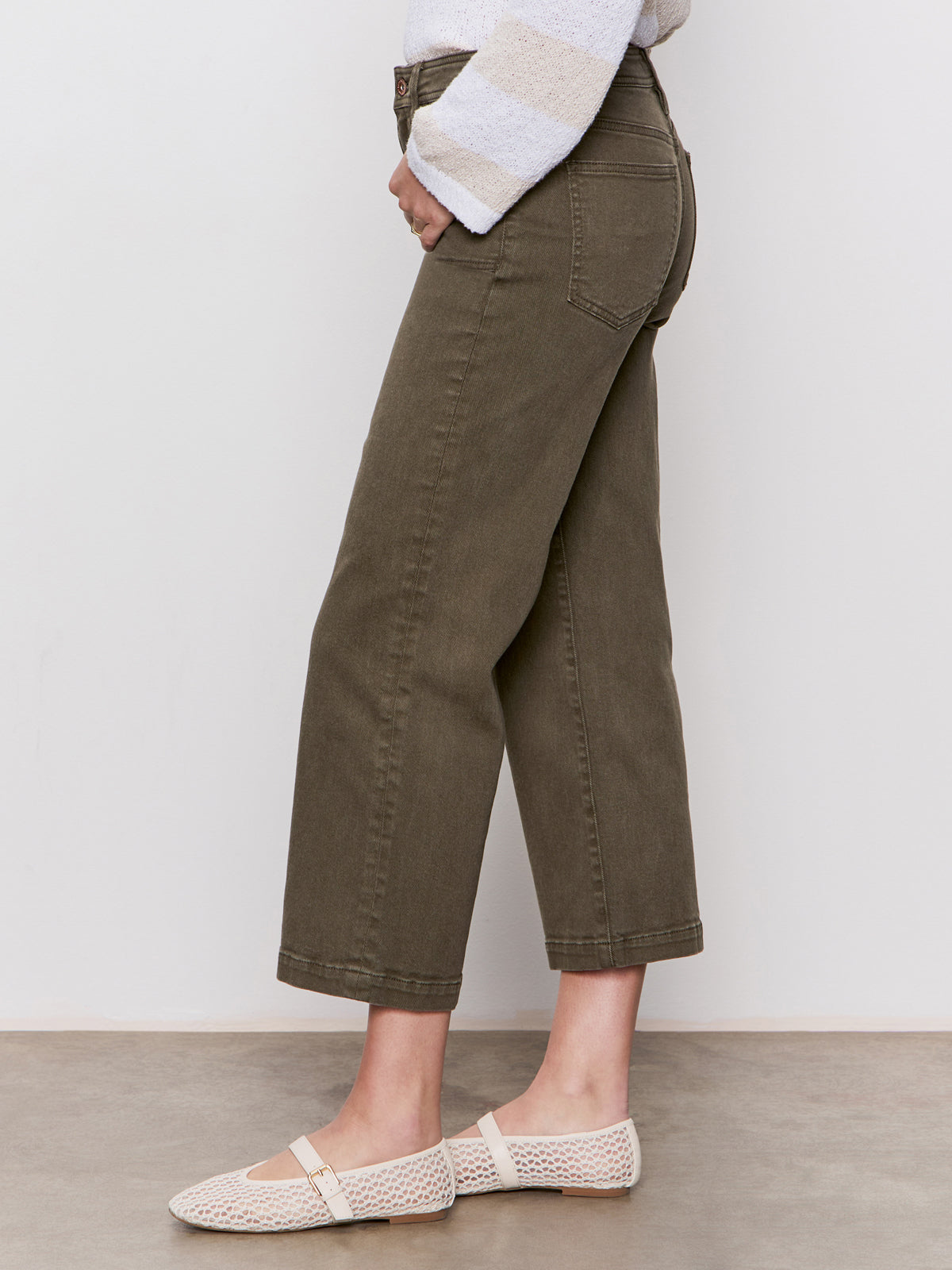A person stands sideways against a plain background, wearing Sanctuary Clothing’s league crop fatigue jeans in olive green, a white and gray striped sweater, and cream-colored woven flats.