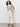 A woman with curly hair wears the Sanctuary Clothing league crop light oat, a sleeveless beige top with black stripes, wide-leg white pants, and black heeled sandals. She stands against a plain white wall with one hand in her pocket.