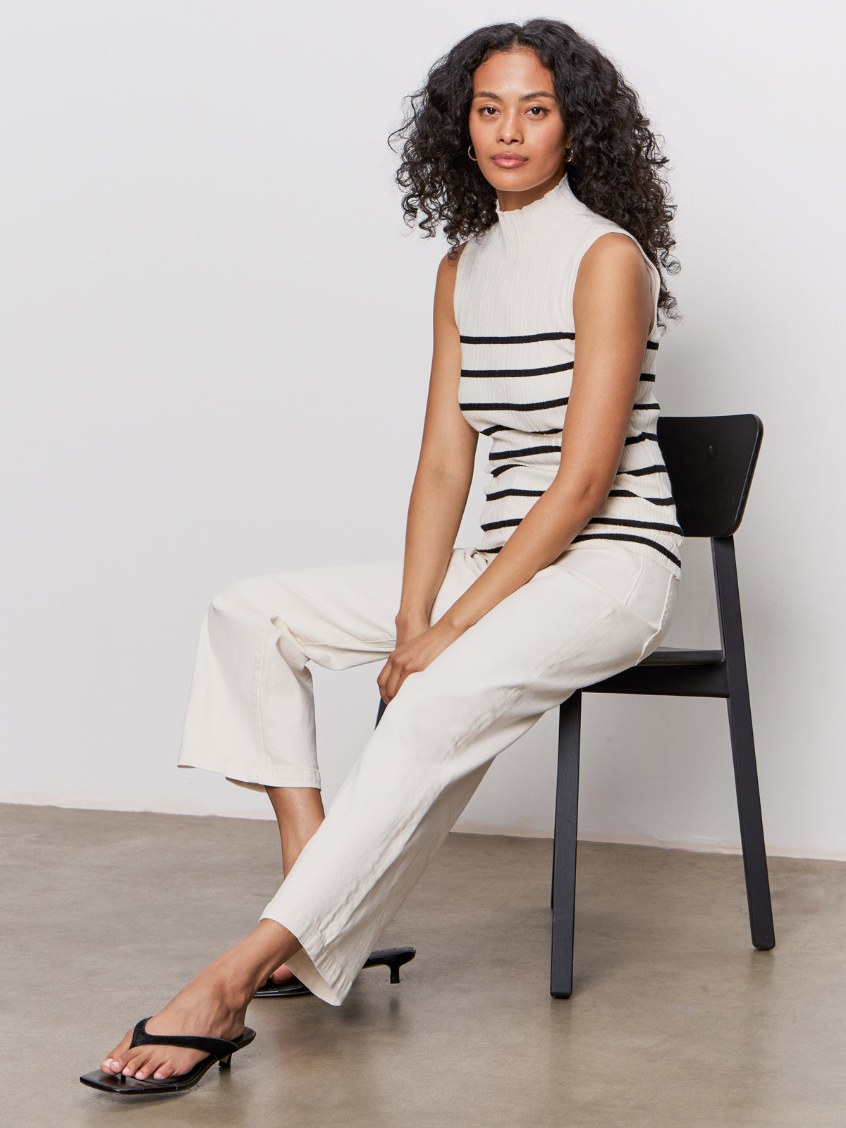 A woman with curly hair sits on a black chair, wearing Sanctuary Clothing’s league crop light oat top with off-white pants and black heeled sandals, against a plain light background.