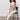 A woman with curly hair sits on a black chair, wearing Sanctuary Clothing’s league crop light oat top with off-white pants and black heeled sandals, against a plain light background.