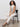 A woman with curly hair sits on a black chair, wearing Sanctuary Clothing’s league crop light oat top with off-white pants and black heeled sandals, against a plain light background.