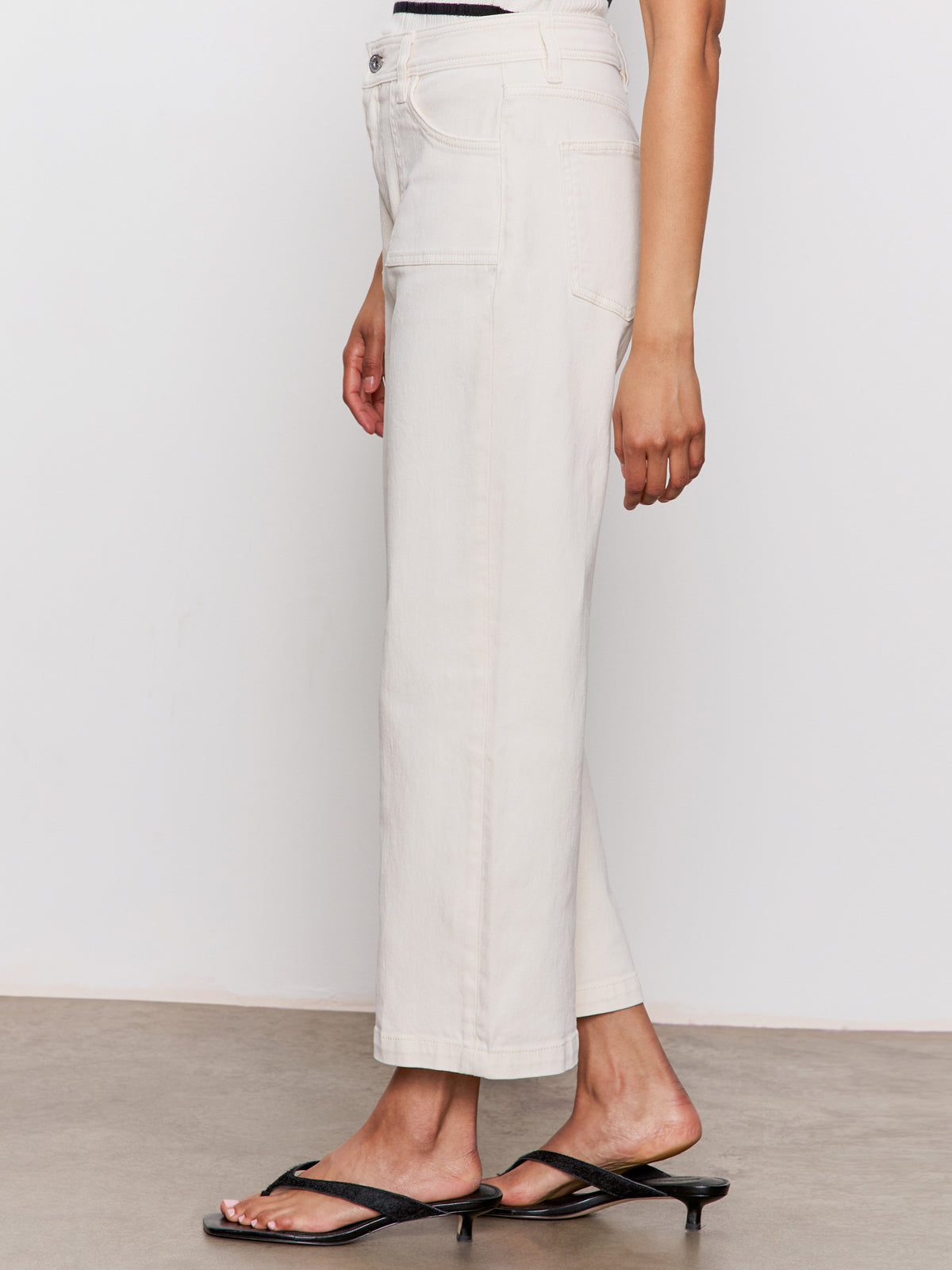 A person models the Sanctuary Clothing league crop light oat jeans, standing sideways in black heeled sandals. Only the lower half of their body is visible against a plain background.