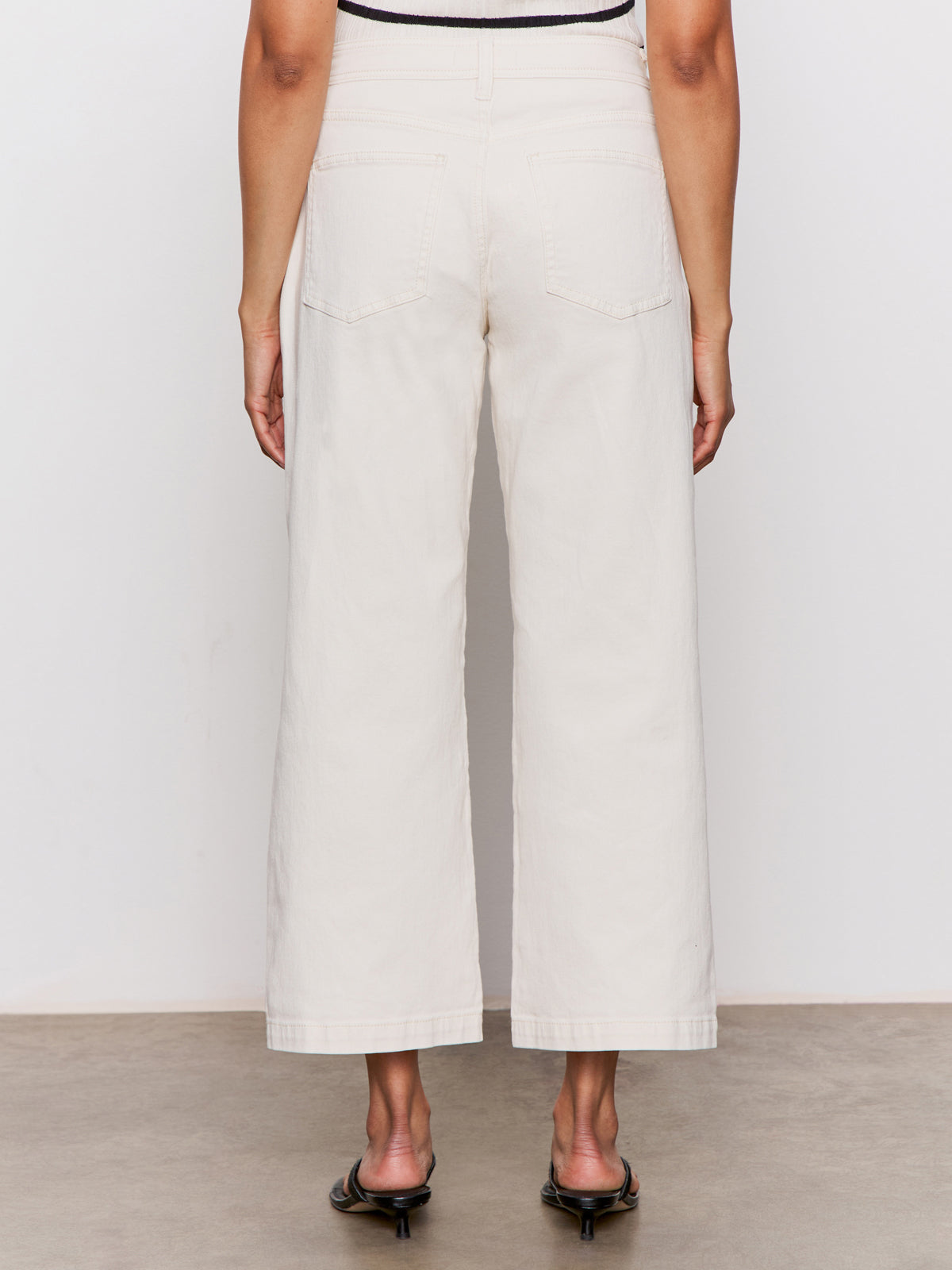 A person stands facing away, wearing Sanctuary Clothing’s league crop light oat high-waisted, wide-leg jeans with back pockets and black heeled sandals against a plain, neutral background.