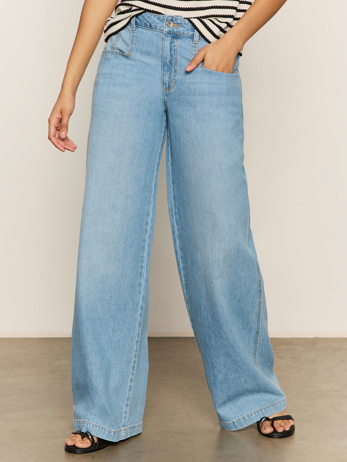 Person wearing Sanctuary Clothing’s Highland Wide Leg Standard Rise Jeans in Sail Away, a black-and-white striped sleeveless top, and black open-toe sandals, standing indoors with one hand in a pocket. Face not visible.