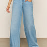 Person wearing Sanctuary Clothing’s Highland Wide Leg Standard Rise Jeans in Sail Away, a black-and-white striped sleeveless top, and black open-toe sandals, standing indoors with one hand in a pocket. Face not visible.