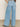 A person wears Sanctuary Clothing's Highland Wide Leg Standard Rise Jeans in Sail Away, paired with a black and white striped top and black sandals, standing with one hand in their back pocket against a plain backdrop.
