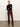 A woman stands against a white wall with arms crossed, wearing a black long-sleeve top, the sculpted brook bootcut pant in chicory by Sanctuary Clothing, and tan heeled boots.