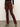 Wearing Sanctuary Clothing's sculpted brook bootcut pant in chicory, paired with a black top and brown pointed-toe heeled boots. The person's head is not visible; they're standing on a gray floor against a plain light wall.