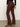 A person stands against a plain wall and concrete floor, facing away with one hand at their side, wearing Sanctuary Clothing’s sculpted brook bootcut pant in chicory, a black long-sleeve top, and tan heeled boots.