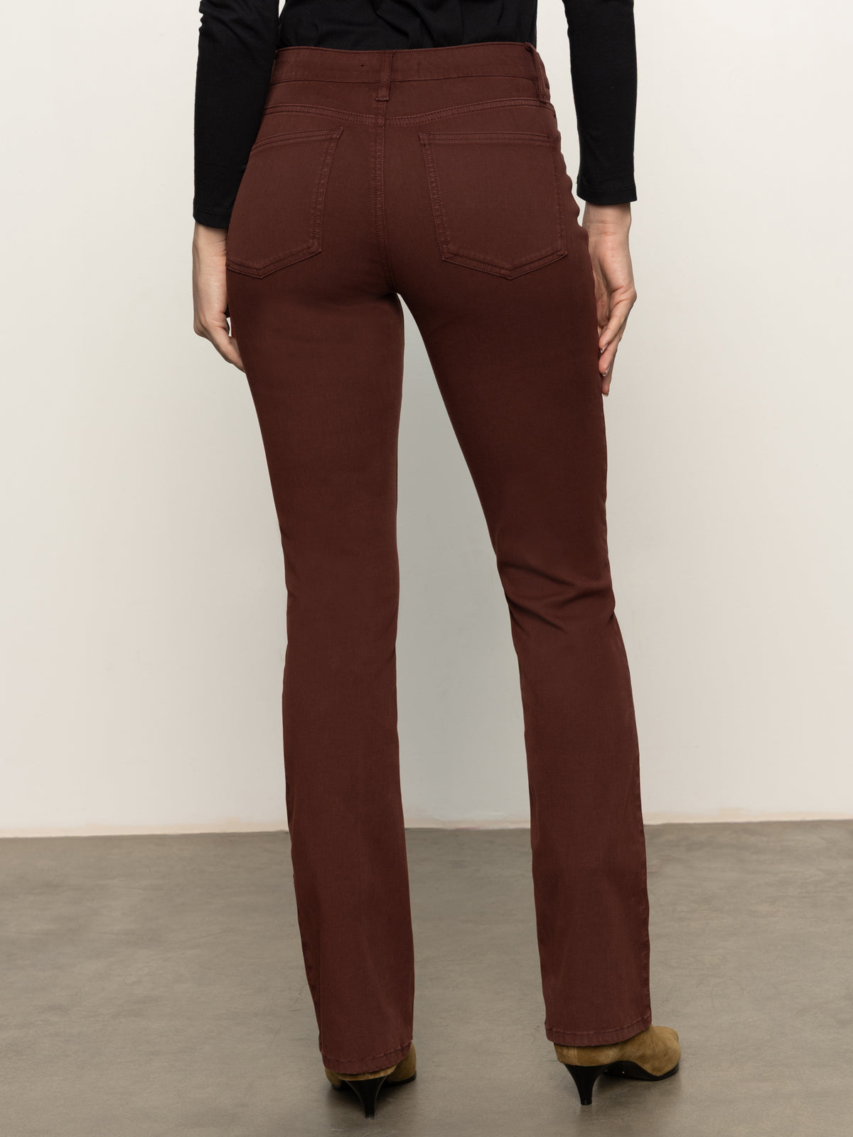 A person wearing Sanctuary Clothing's sculpted brook bootcut pant in chicory faces away from the camera, paired with a black long-sleeve top on a gray floor against a plain light background.