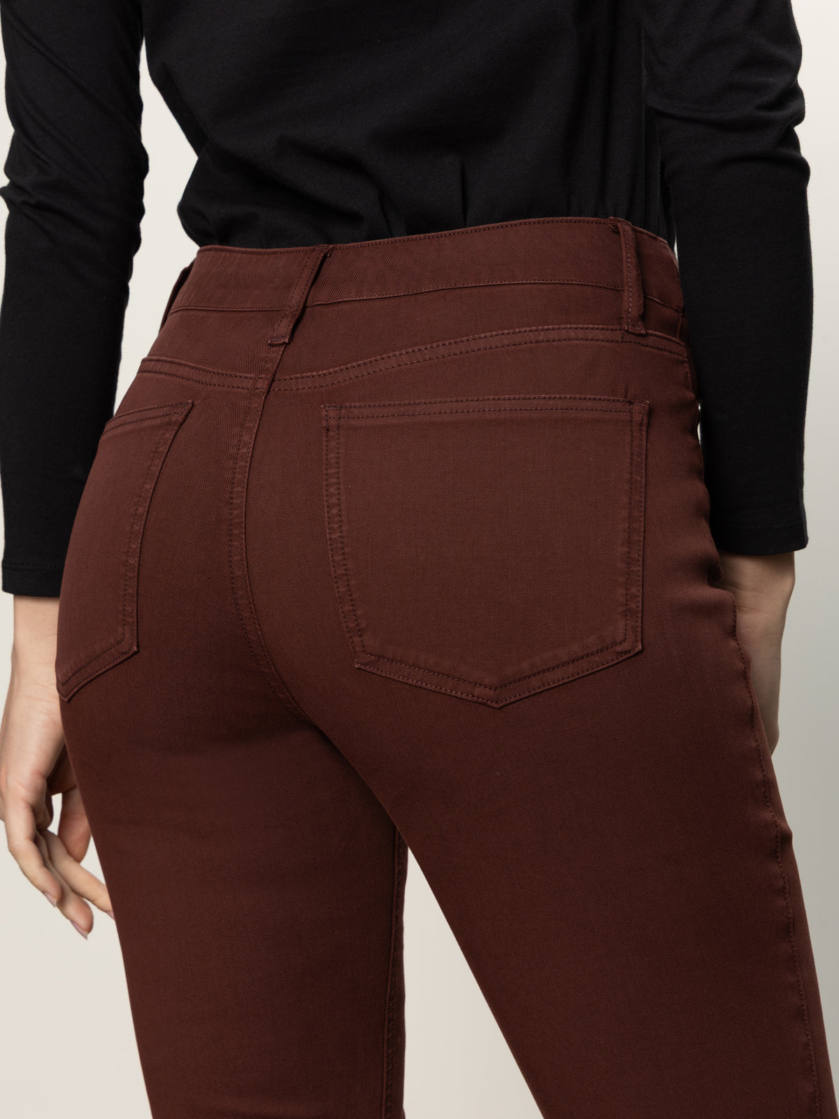 A person wearing Sanctuary Clothing’s sculpted brook bootcut pant in chicory, a high-waisted dark brown jean with back pockets, paired with a black long-sleeve top—shown from the waist down and photographed from behind.