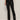 Wearing Sanctuary Clothing's slick brook bootcut pant in black, paired with black pointed-toe boots, a person stands against a neutral background with one hand in their pocket and their upper body mostly out of frame.
