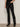 Wearing Sanctuary Clothing's slick brook bootcut pant in black, paired with black pointed-toe boots, a person stands against a neutral background with one hand in their pocket and their upper body mostly out of frame.
