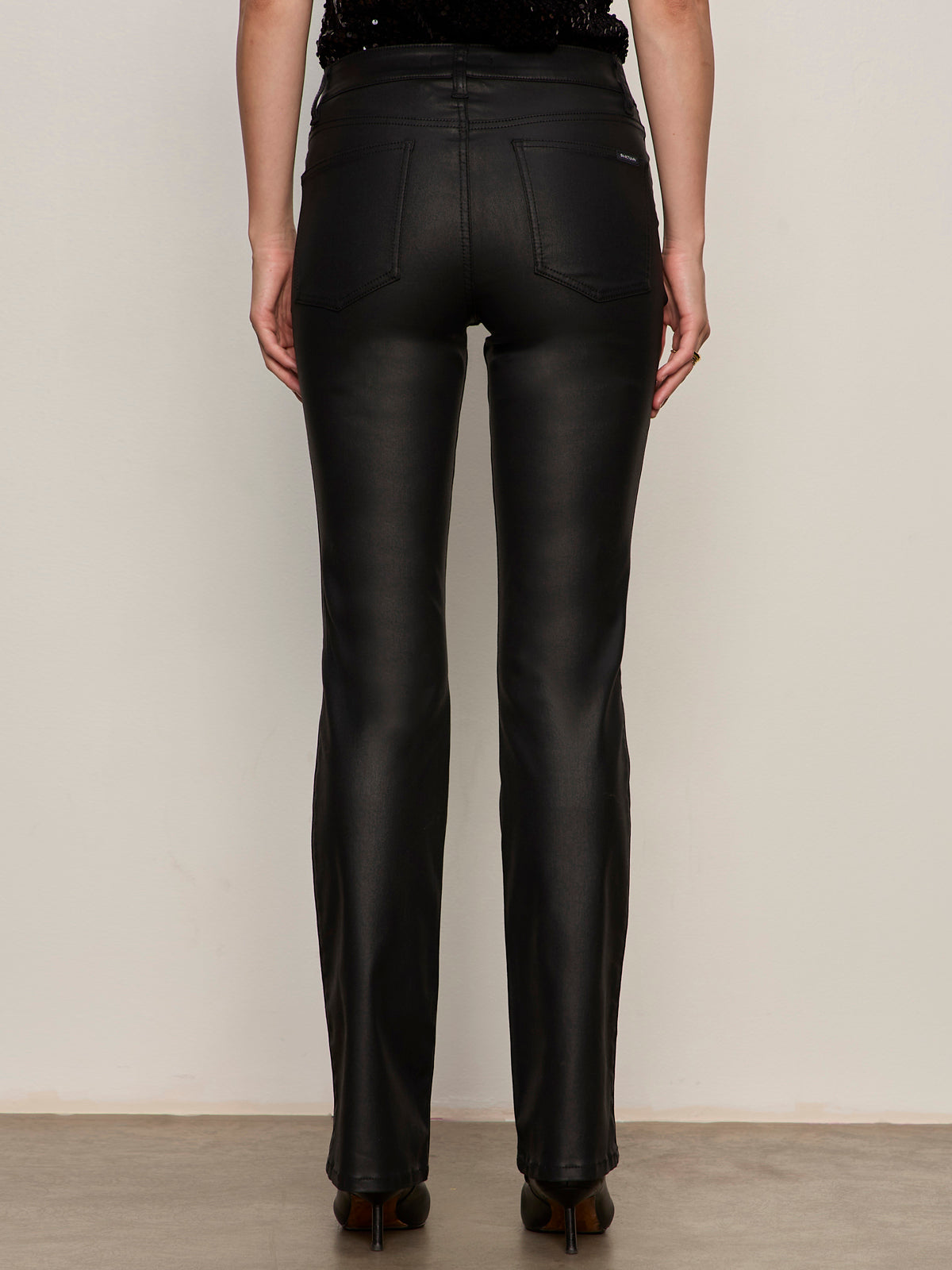 A person faces away from the camera, wearing Sanctuary Clothing's slick brook bootcut pant in black—high-waisted, straight-leg, shiny material—paired with dark heeled shoes against a plain neutral background.