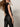 A woman with long brown hair, sunglasses, and a black sequin top wears Sanctuary Clothing's slick brook bootcut pant in black, turning slightly to the side against a plain light background.