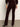 A person stands on a gray floor facing away, wearing the Sanctuary Clothing velveteen brook bootcut pant in dark crimson, paired with a matching top and heeled ankle boots. Their left arm hangs by their side while the right is bent.