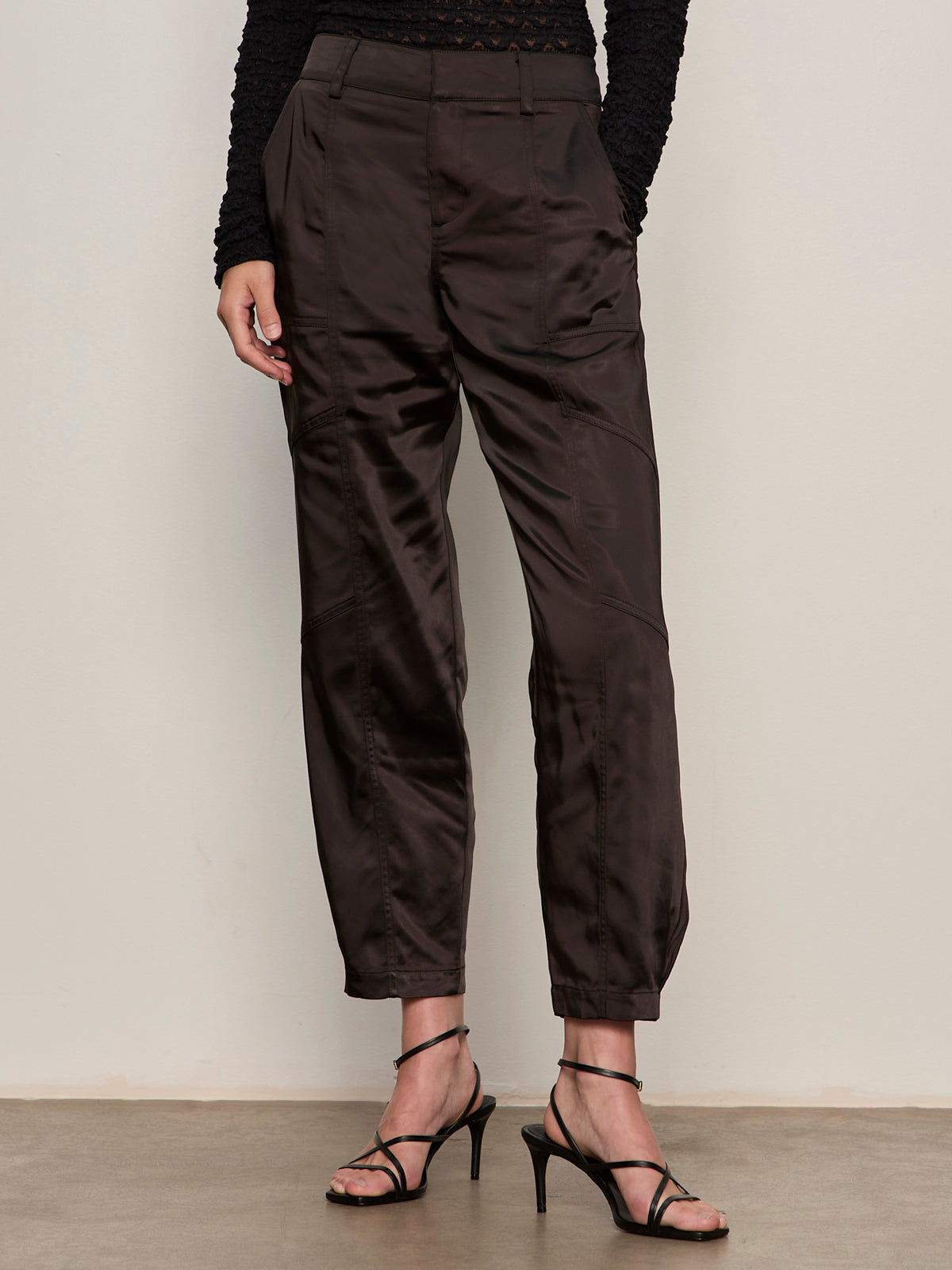 A person wearing Sanctuary Clothing's sahara satin tapered pant in black, paired with a long-sleeved black lace top and black strappy high-heeled sandals, stands against a plain light-colored wall.
