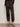 A person wearing Sanctuary Clothing's sahara satin tapered pant in black, paired with a long-sleeved black lace top and black strappy high-heeled sandals, stands against a plain light-colored wall.