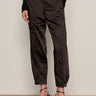 A person wearing Sanctuary Clothing's sahara satin tapered pant in black, paired with a long-sleeved black lace top and black strappy high-heeled sandals, stands against a plain light-colored wall.